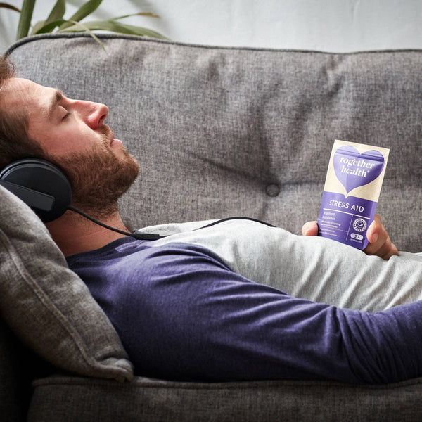 Mann liegt auf Sofa, hört mit geschlossenen Augen Musik und hält eine Tüte Together Health Stress Aid in der Hand