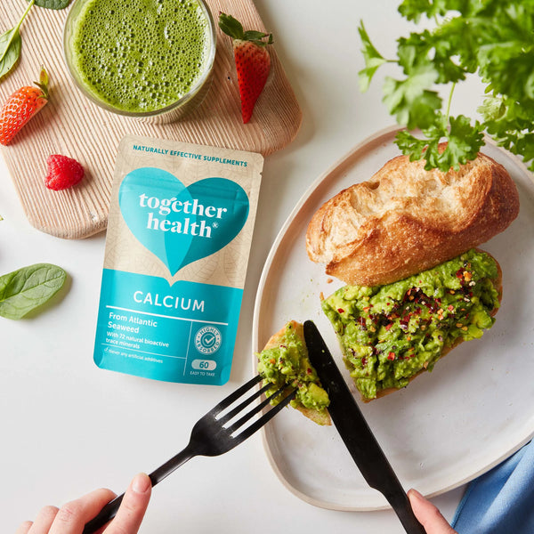 zwei Hände schneiden mit Messer und Gabel ein Brötchen mit Gemüsepaste, daneben liegt die Together Health Tüte veganes Calcium