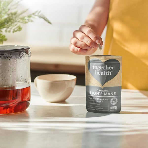 Eine Hand nimmt eine Kapsel aus einer Tüte Together Health Organic Lion's Mane, die auf einem Tisch neben einer Teetasse und einer Teekanne steht