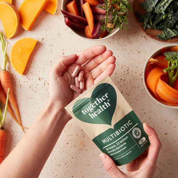 Hand zeigt über einem Tisch Kapseln von Together Health Multibiotic und ringsherum stehen Schalen mit frischen Gemüsestücken