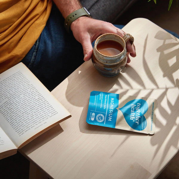 Hand hält eine Kaffeetasse auf einem Tischchen, daneben liegen die Together Health Magnesium Tüte und ein Buch“