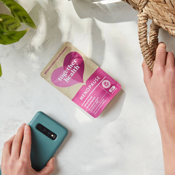 Auf weisser Tischdecke liegt eine Hand mit Handy, andere Frauenhand hält einen Korb, dazwischen liegt eine Tüte Together Health Menopause
